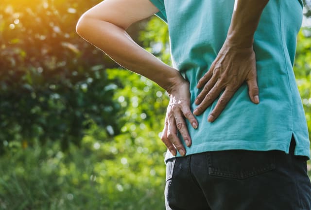 A person standing outdoors with one hand on their lower back and the other on their hip, indicating back pain